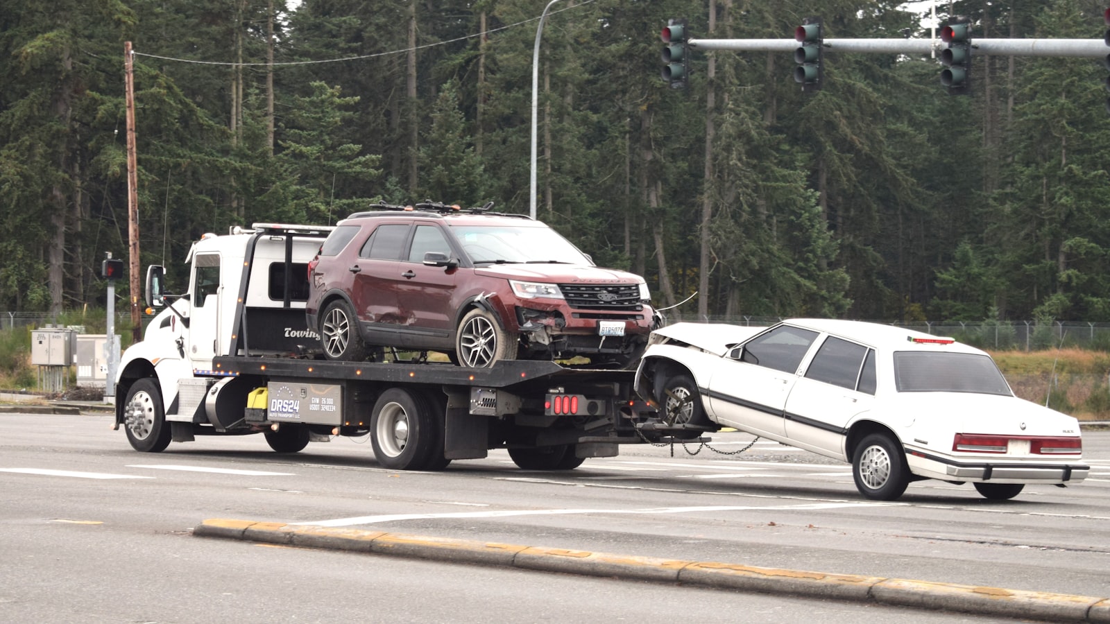 Flatbed tow truck hauling a damaged SUV while towing a sedan away from a crash scene