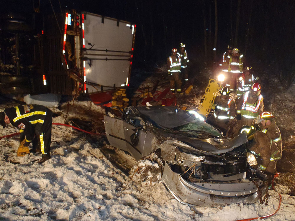 Night crash scene with a wrecked car crushed against an overturned semi-trailer and firefighters working under floodlights