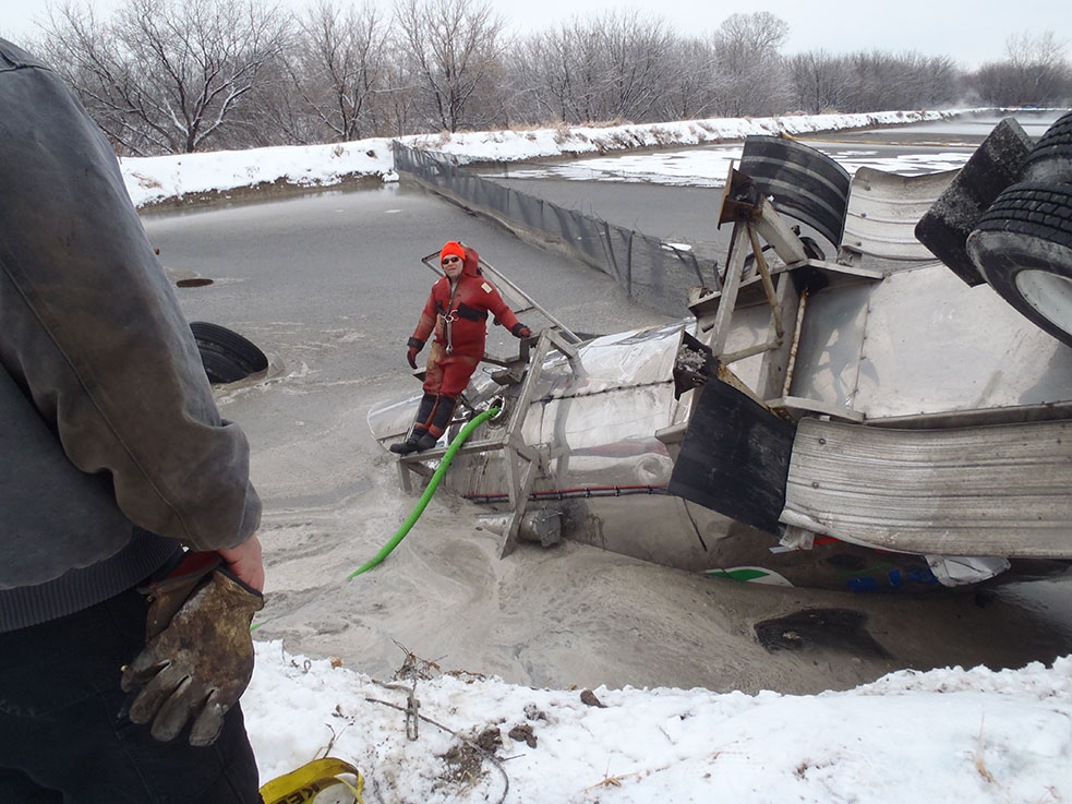 Overturned tanker trailer lying in a frozen lagoon while a worker in a dry suit pumps out its load