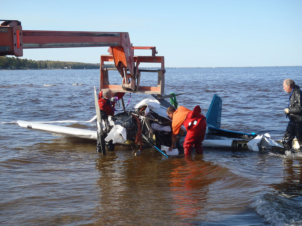 Small aircraft lifted from the bay on chains by a boom crane with the crew standing in the water
