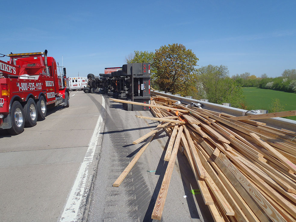 Mike's Wrecker heavy wrecker on a highway beside an overturned truck and its spilled load of lumber